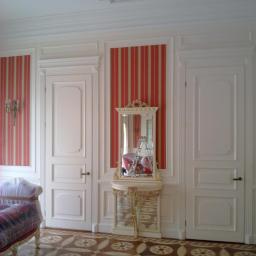 Drewkomplet Spółka z Ograniczoną Odpowiedzialnością - Eleganckie wnętrze z dwiema białymi drzwiami i lustrem na ozdobnej konsoli, wzorzysta podłoga i ściany w czerwono-beżowe pasy. Klasyczny design.