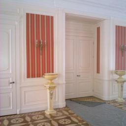 Drewkomplet Spółka z Ograniczoną Odpowiedzialnością - Klasyczne, białe drzwi wewnętrzne z dekoracyjnymi panelami i czerwono-białą tapetą w paski, uzupełnione o zdobione, kremowe postumenty i wzorzystą podłogę.