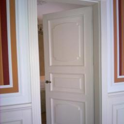 Drewkomplet Spółka z Ograniczoną Odpowiedzialnością - Otwarta, biała, płycinowa drzwi wewnętrzne z ozdobną ościeżnicą, widok na wnętrze pokoju. Dekoracyjne pasy na ścianach po bokach drzwi.