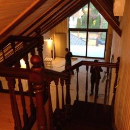 Drewkomplet Spółka z Ograniczoną Odpowiedzialnością - Wnętrze poddasza z drewnianymi schodami i balustradą w klasycznym stylu. Widoczny fragment okna i osoby pracujące w tle. Ciepłe oświetlenie.