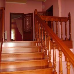 Drewkomplet Spółka z Ograniczoną Odpowiedzialnością - Polerowane, drewniane schody z ozdobną balustradą, prowadzące w górę. W tle widoczne drzwi z dekoracyjnym szkłem i zasłoną, całość w ciepłej, różowej tonacji.