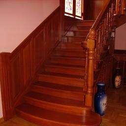 Drewkomplet Spółka z Ograniczoną Odpowiedzialnością - Klasyczne schody drewniane z ozdobnymi balustradami i stopniami w ciepłym odcieniu brązu, na tle różowej ściany i parkietu. Widoczne wazony dekoracyjne.