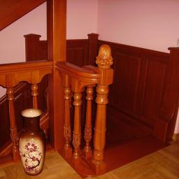 Drewkomplet Spółka z Ograniczoną Odpowiedzialnością - Drewniane schody z rzeźbioną balustradą, widok z dołu. Na pierwszym planie wazon z motywem kwiatowym. Ściany w jasnym odcieniu różu. Ciepłe oświetlenie podkreśla detale.
