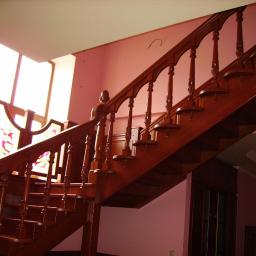 Drewkomplet Spółka z Ograniczoną Odpowiedzialnością - Klasyczne schody drewniane z ozdobnymi balustradami i tralkami, oświetlone naturalnym światłem wpadającym przez okno ze zdobioną szybą.