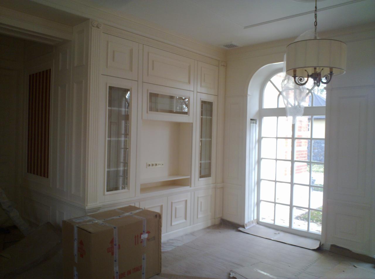 Elegancka zabudowa meblowa w kremowym odcieniu, z przeszklonymi witrynami i półkami, w jasnym wnętrzu z dużym oknem łukowym. Karton na podłodze sugeruje świeży montaż.