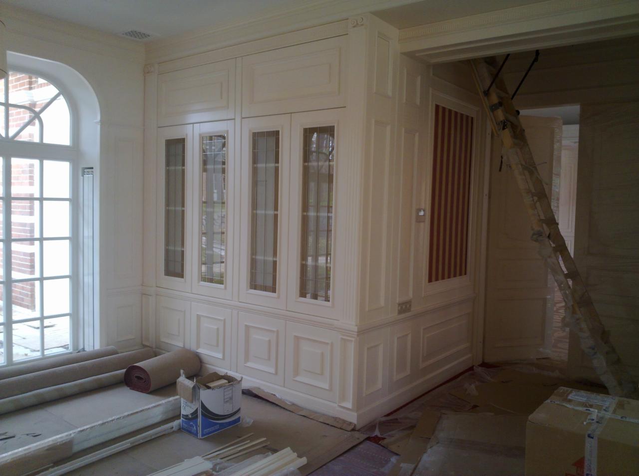 Klasyczna zabudowa meblowa z przeszklonymi witrynami i dekoracyjną sztukaterią w jasnym wnętrzu z elementami wykończeniowymi i drabiną.