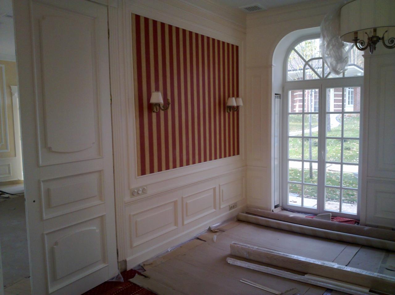 Klasyczne wnętrze z otwartymi drzwiami, ścianą w pasy i dużym oknem. Widoczne listwy przypodłogowe i elementy dekoracyjne. Podłoga zabezpieczona folią.