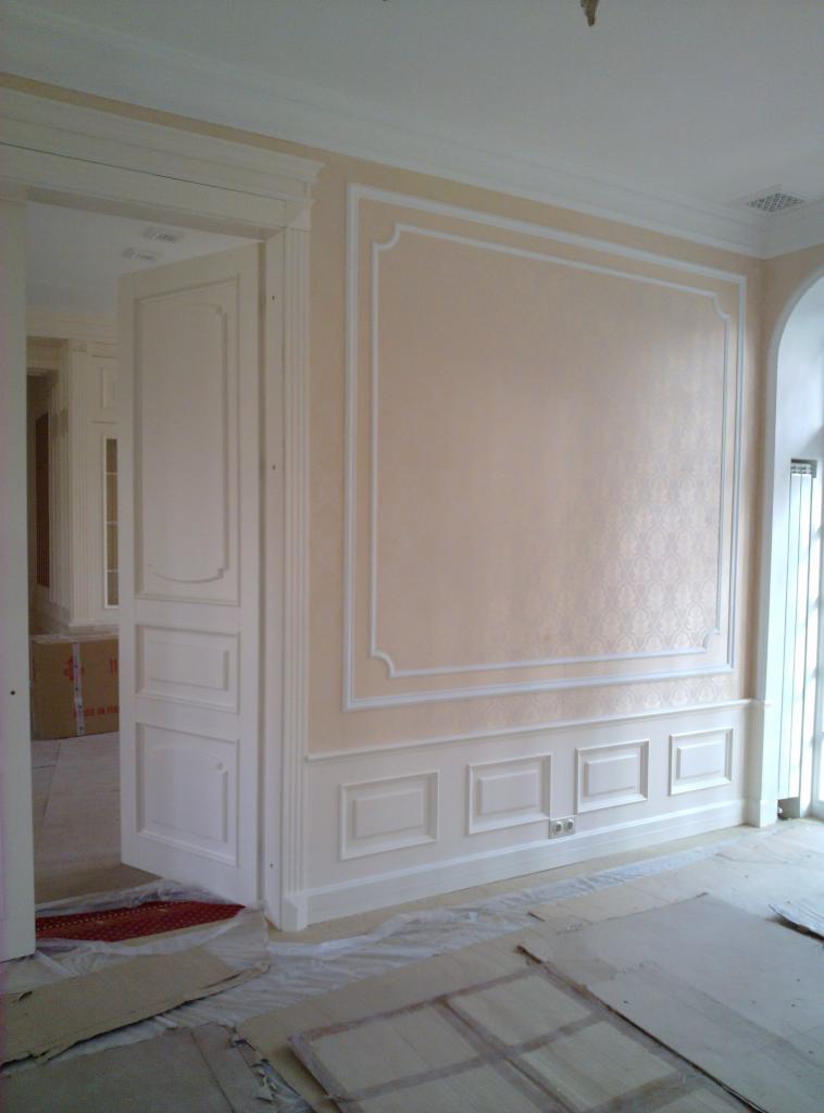 Eleganckie, beżowe ściany z dekoracyjnymi, białymi listwami i panelami. Widoczne otwarte drzwi z białymi płycinami. Podłoga zabezpieczona folią malarską.