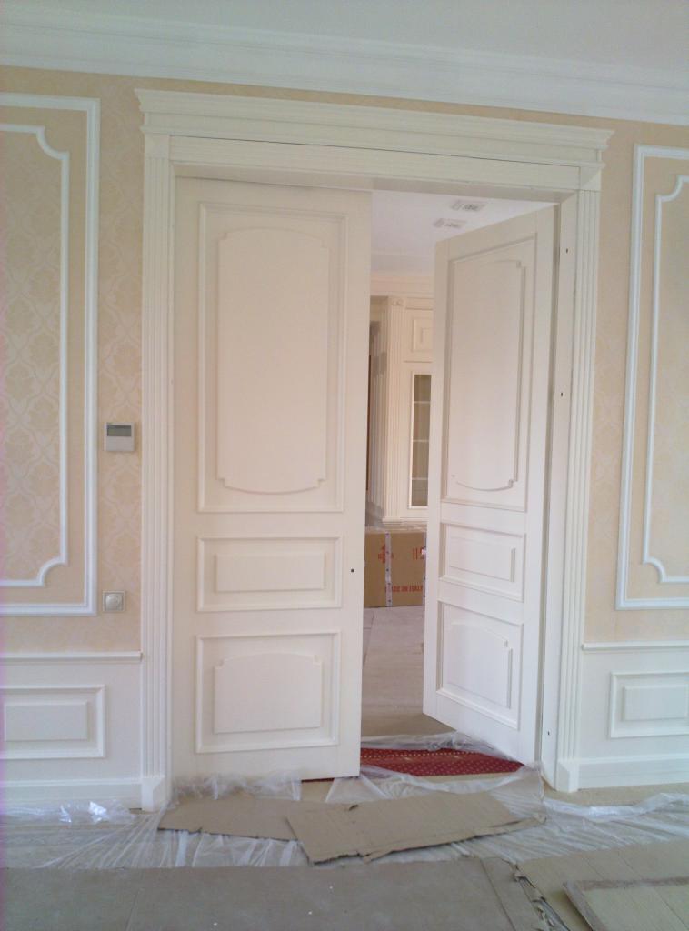 Otwarty, dwuskrzydłowy portal drzwiowy z dekoracyjnymi listwami w kremowym odcieniu. Widoczne zabezpieczenia podłoża. W tle pomieszczenie z zabudową meblową.