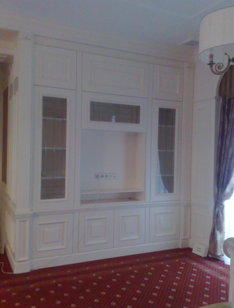 Biała, zabudowana szafa wnękowa z przeszklonymi witrynami i miejscem na telewizor, wkomponowana w klasyczne wnętrze z czerwonym dywanem.