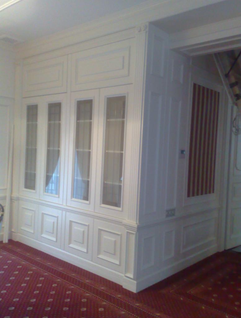 Biała, zabudowana szafa z przeszklonymi drzwiami, wkomponowana w ścianę z dekoracyjnymi panelami. Czerwony dywan z wzorem na podłodze, klasyczny wystrój.