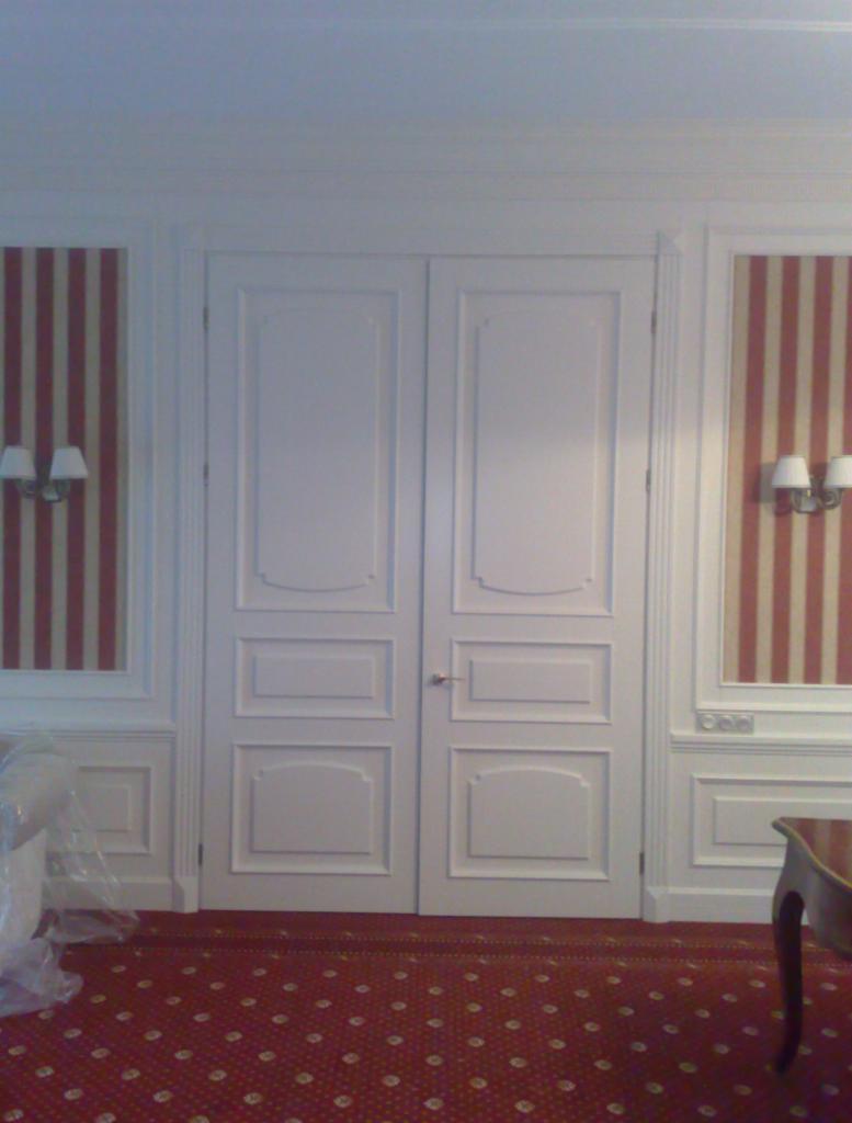 Eleganckie, białe dwuskrzydłowe drzwi wewnętrzne z dekoracyjnymi panelami, wkomponowane w ścianę z tapetą w czerwono-białe pasy i ozdobną sztukaterią.