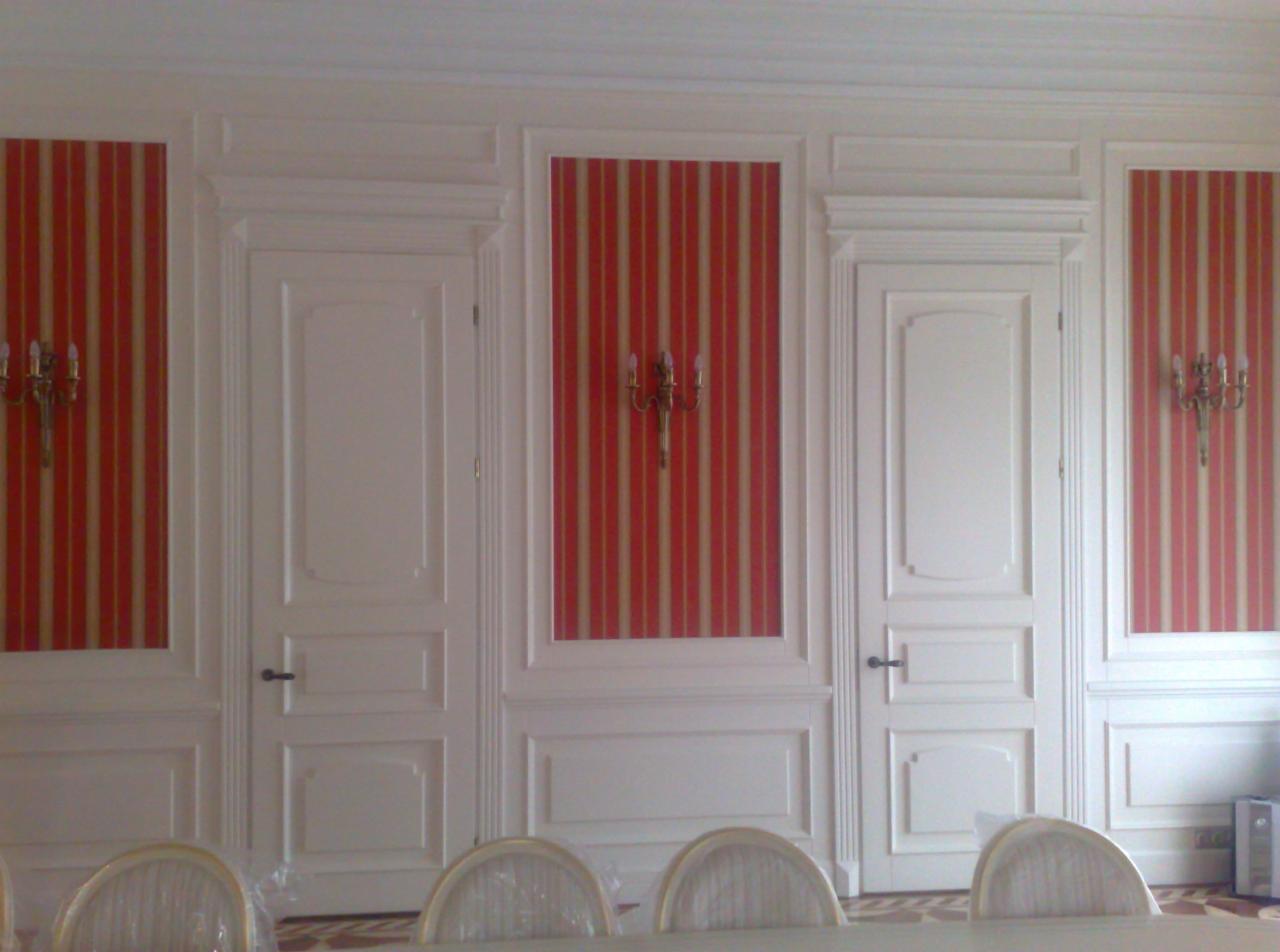 Eleganckie, białe drzwi wewnętrzne z dekoracyjnymi panelami ściennymi w paski, oświetlone kinkietami. Widoczne krzesła w dolnej części kadru.