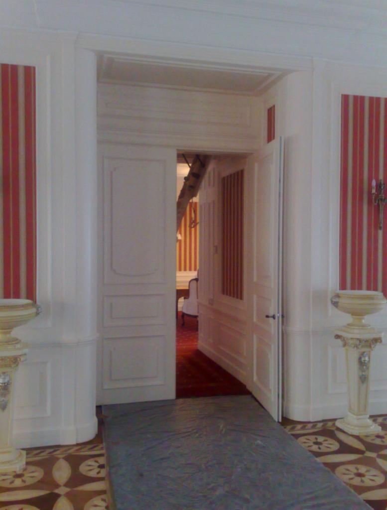 Otwarta, biała, dwuskrzydłowa, płycinowa  drzwi wewnętrzne z widokiem na pokój z tapetami w pasy. Podłoga zabezpieczona folią malarską.