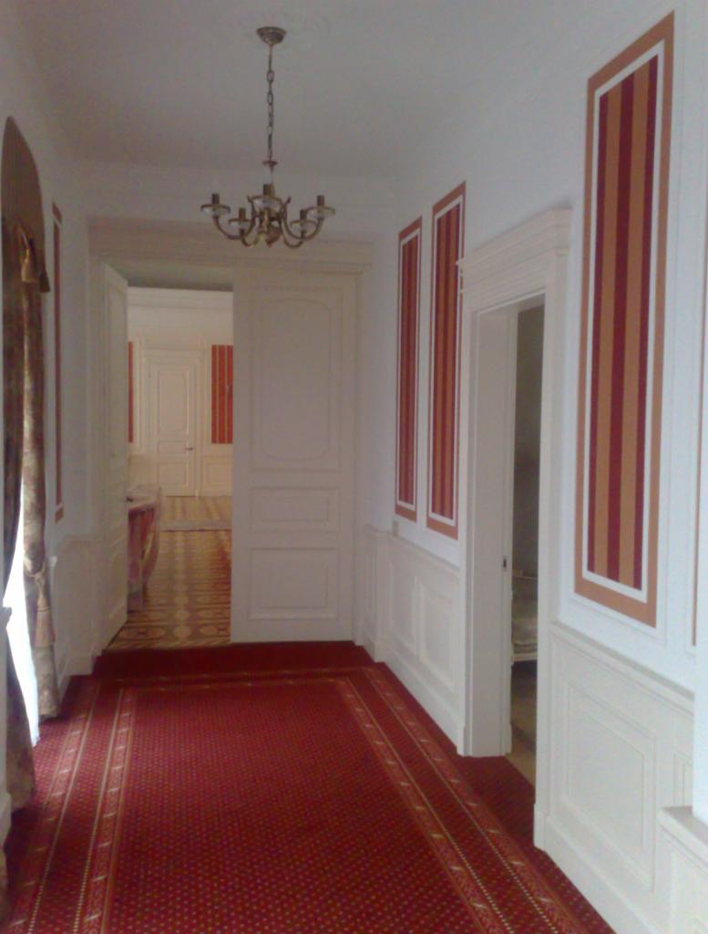 Elegancki korytarz z czerwonym dywanem i białymi panelami ściennymi. Widoczne drzwi wewnętrzne i ozdobne malowanie pionowych pasów na ścianach.