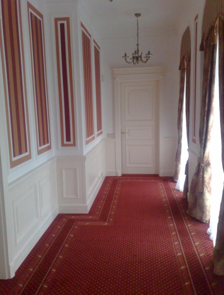 Korytarz z czerwonym dywanem, białymi panelami ściennymi z bordowo-złotymi pasami i klasycznymi, białymi drzwiami z ościeżnicą. Nad drzwiami wisi żyrandol.