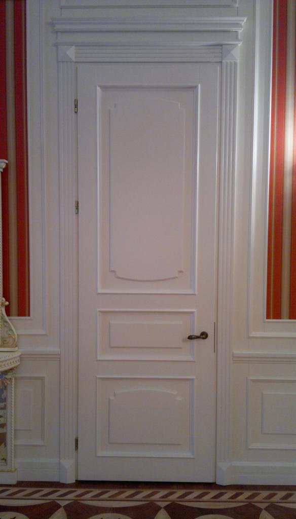 Eleganckie, białe drzwi wewnętrzne z dekoracyjnymi panelami, dopasowane do klasycznego wystroju wnętrza z tapetą w paski i ozdobną podłogą.