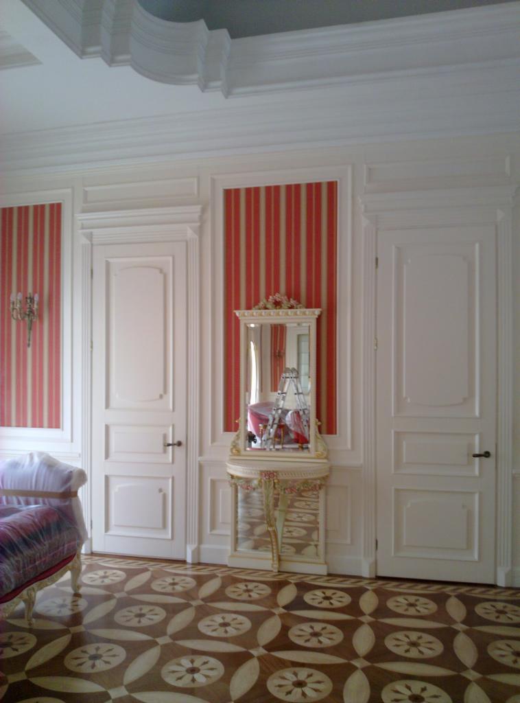 Eleganckie wnętrze z dwiema białymi drzwiami i lustrem na ozdobnej konsoli, wzorzysta podłoga i ściany w czerwono-beżowe pasy. Klasyczny design.