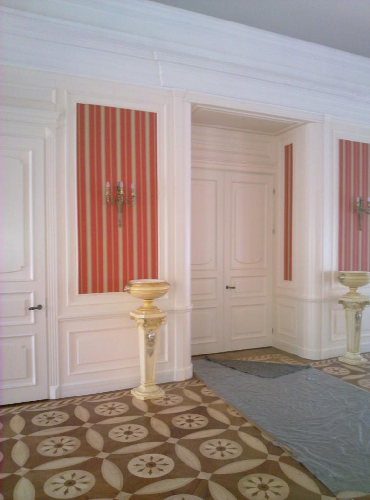 Klasyczne, białe drzwi wewnętrzne z dekoracyjnymi panelami i czerwono-białą tapetą w paski, uzupełnione o zdobione, kremowe postumenty i wzorzystą podłogę.