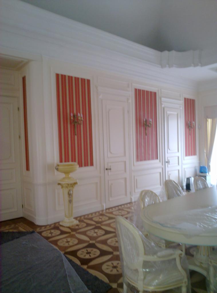 Elegancka aranżacja wnętrza z białymi, klasycznymi drzwiami i panelami ściennymi w paski, uzupełniona o zdobione kinkiety i dekoracyjną kolumnę.