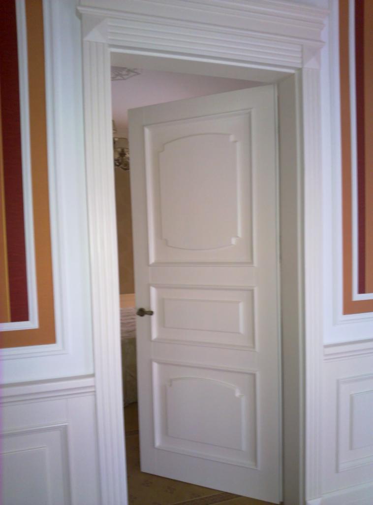 Otwarta, biała, płycinowa drzwi wewnętrzne z ozdobną ościeżnicą, widok na wnętrze pokoju. Dekoracyjne pasy na ścianach po bokach drzwi.