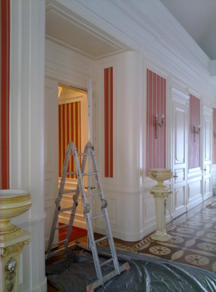 Korytarz w trakcie renowacji: białe ściany z czerwonymi pasami, drabina, zdobione kolumny i podłoga. Widoczne detale architektoniczne i etap prac wykończeniowych.