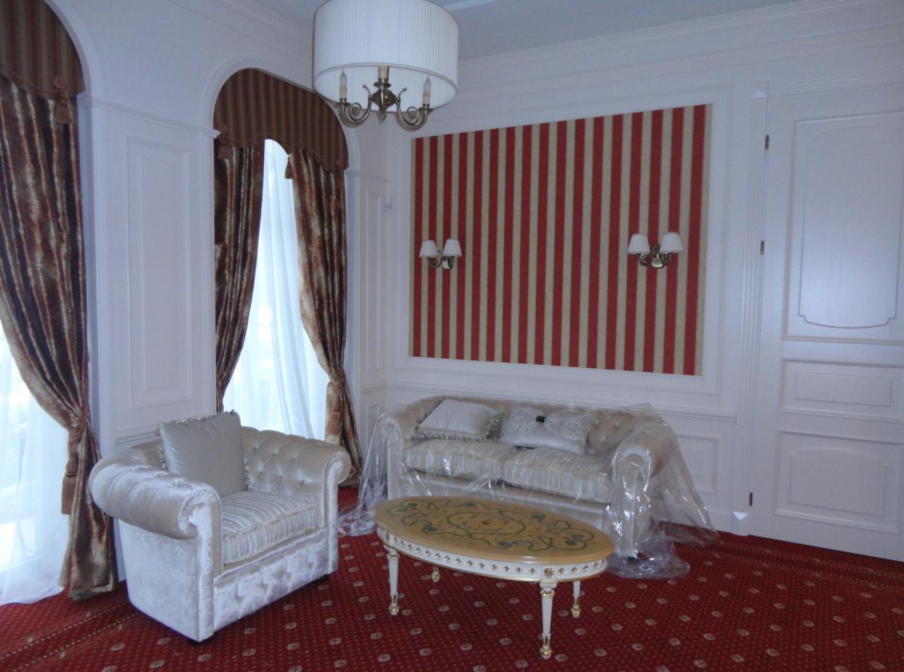 Elegancki salon z beżowym fotelem, sofą pod folią ochronną i owalnym stolikiem kawowym na czerwonym dywanie. Okno z dekoracyjnymi zasłonami i stylowe oświetlenie.