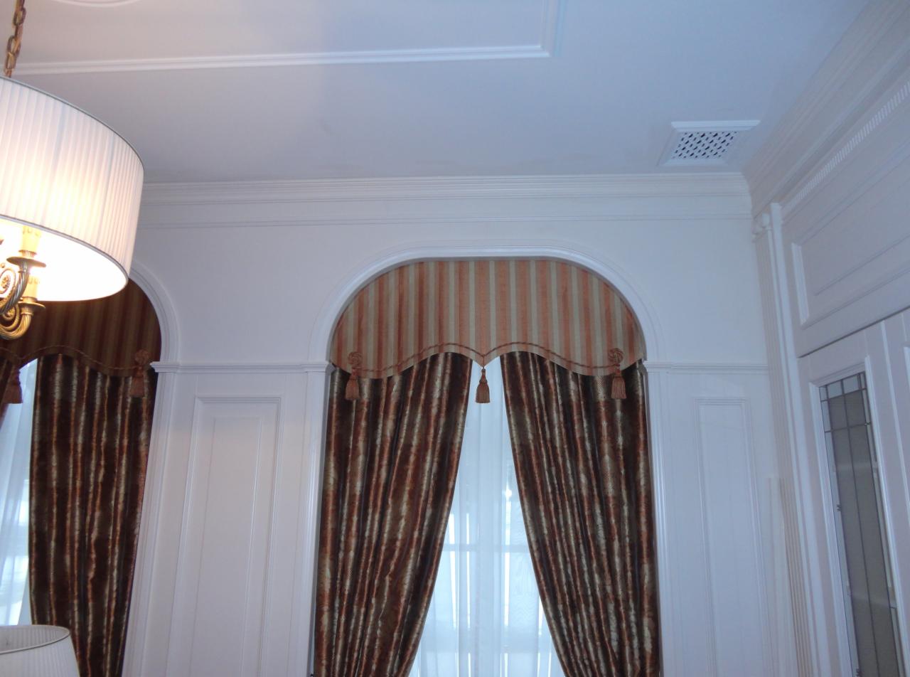 Eleganckie okno z dekoracyjnymi, brązowo-złotymi zasłonami w paski, zwieńczone łukowym lambrekinem z frędzlami, w klasycznym, białym wnętrzu.