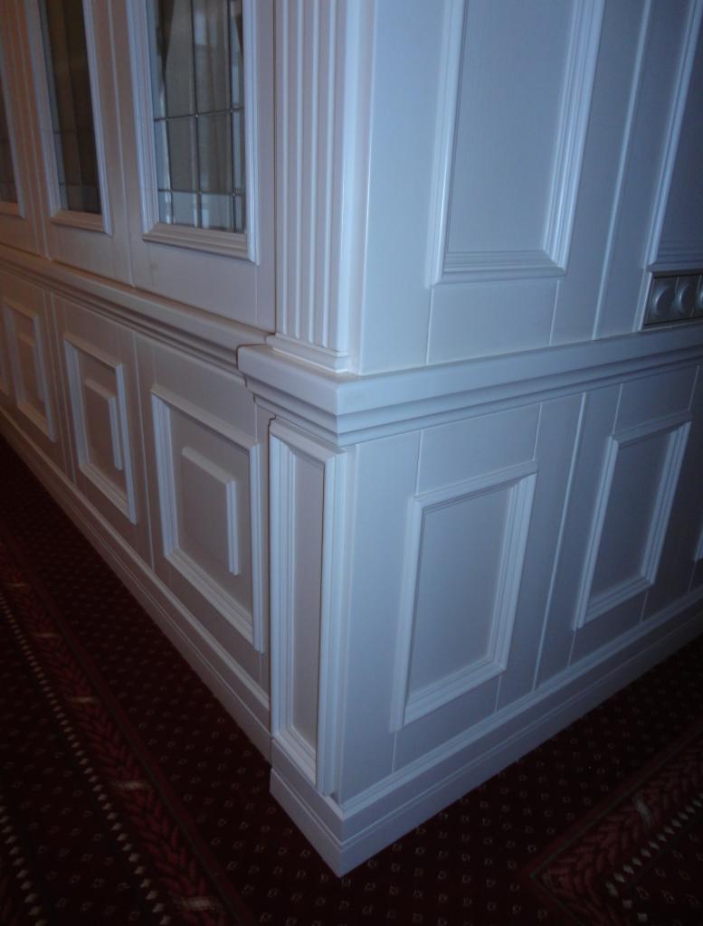 Biała boazeria angielska z frezowanymi panelami i przeszklonymi szafkami, zamontowana w rogu pomieszczenia z wzorzystą wykładziną. Klasyczny design.