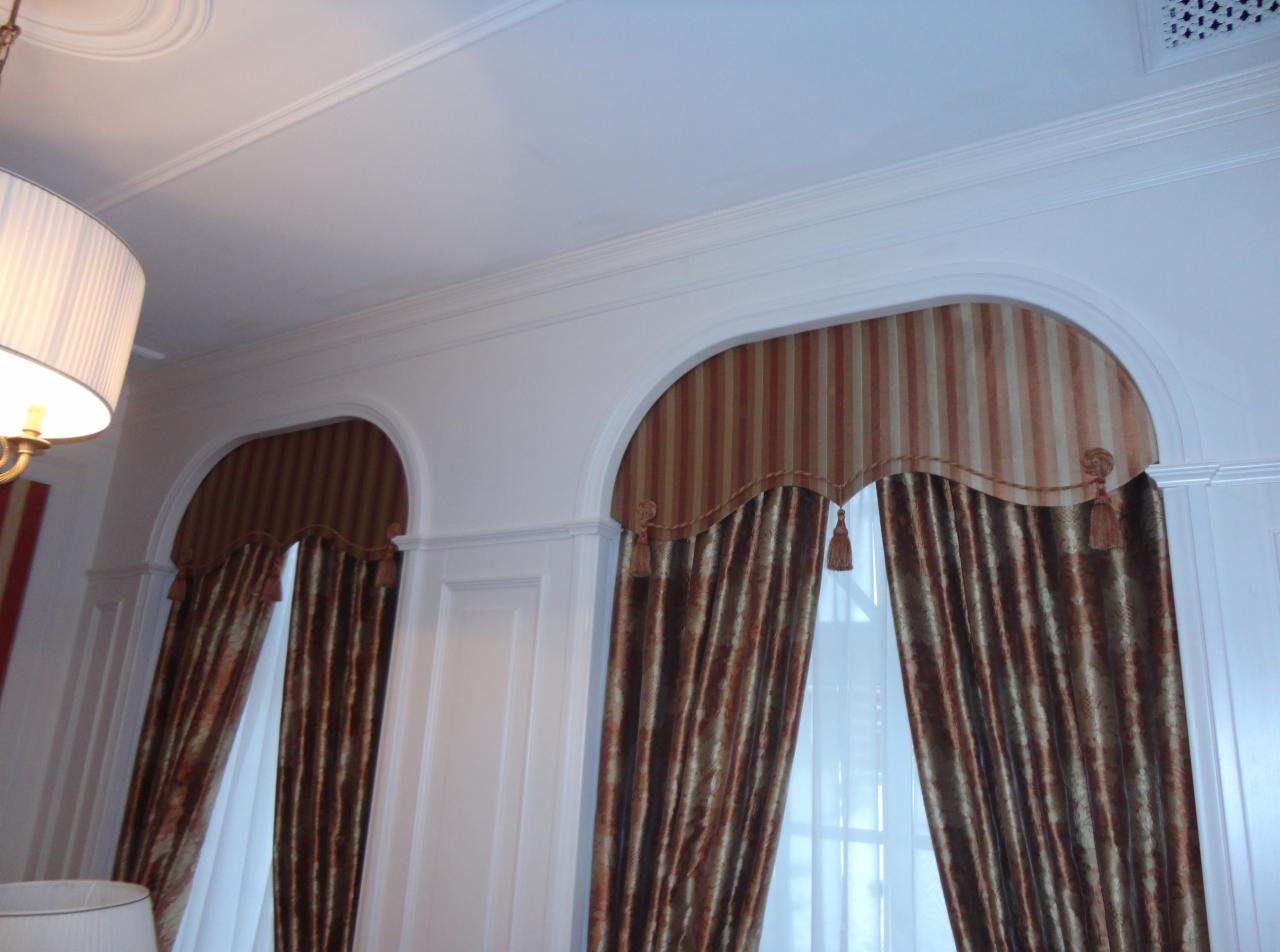 Dwa okna z łukowatymi nadprożami, udekorowane brązowo-złotymi, pionowo prążkowanymi zasłonami z dekoracyjnymi chwostami. Białe ściany i sufit z listwami.