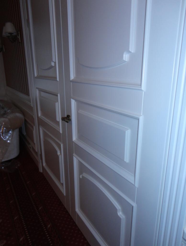 Białe, dwuskrzydłowe drzwi wewnętrzne z dekoracyjnymi frezowaniami i mosiężną klamką, widoczne z boku na tle wzorzystej ściany i dywanu.