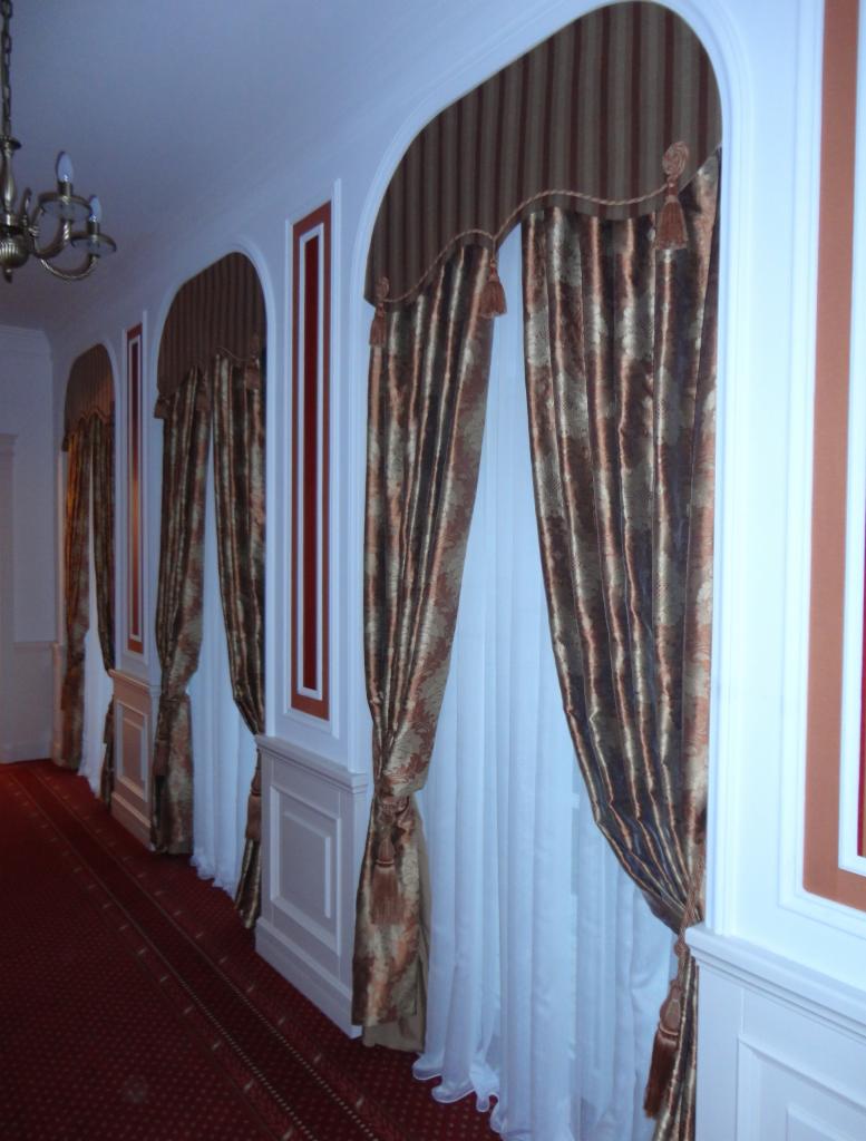 Eleganckie, brązowo-złote zasłony z frędzlami, upięte po bokach okien w łukowatych wnękach, w korytarzu z czerwonym dywanem i białymi panelami ściennymi.