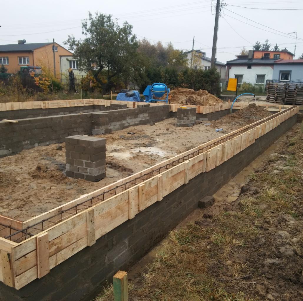 Wykop pod fundamenty z częściowo wylanymi ścianami, drewnianym szalunkiem i zbrojeniem, na placu budowy widoczna betoniarka i materiały budowlane.