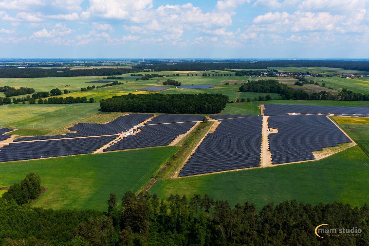 Rozległa farma paneli słonecznych wkomponowana w zielony krajobraz pól i lasów, widok z lotu ptaka, błękitne niebo z lekkimi chmurami w tle.