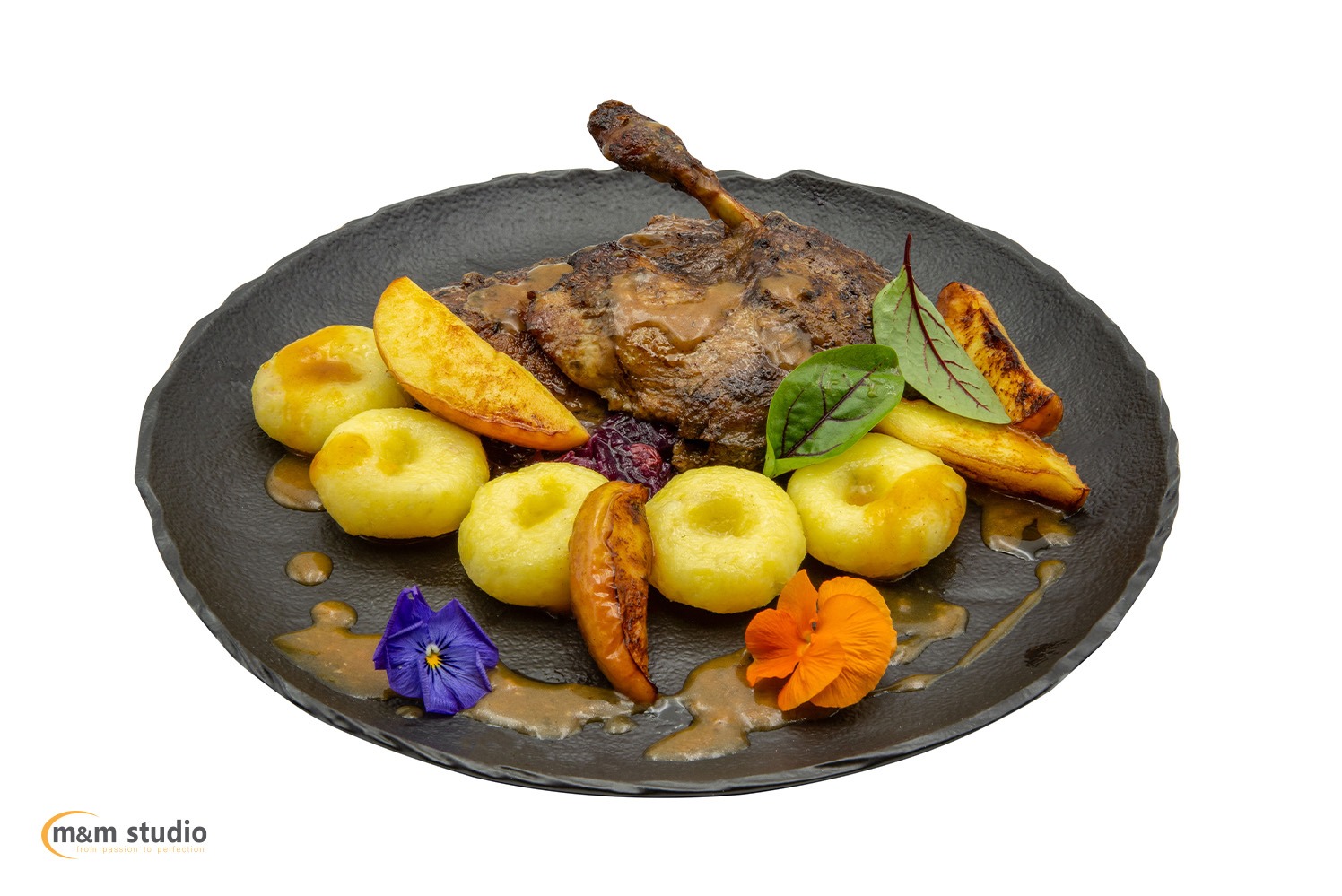 Apetyczna noga gęsi z kluskami i pieczonymi jabłkami na czarnym talerzu, udekorowana jadalnymi kwiatami. Profesjonalna fotografia kulinarna z bliska.