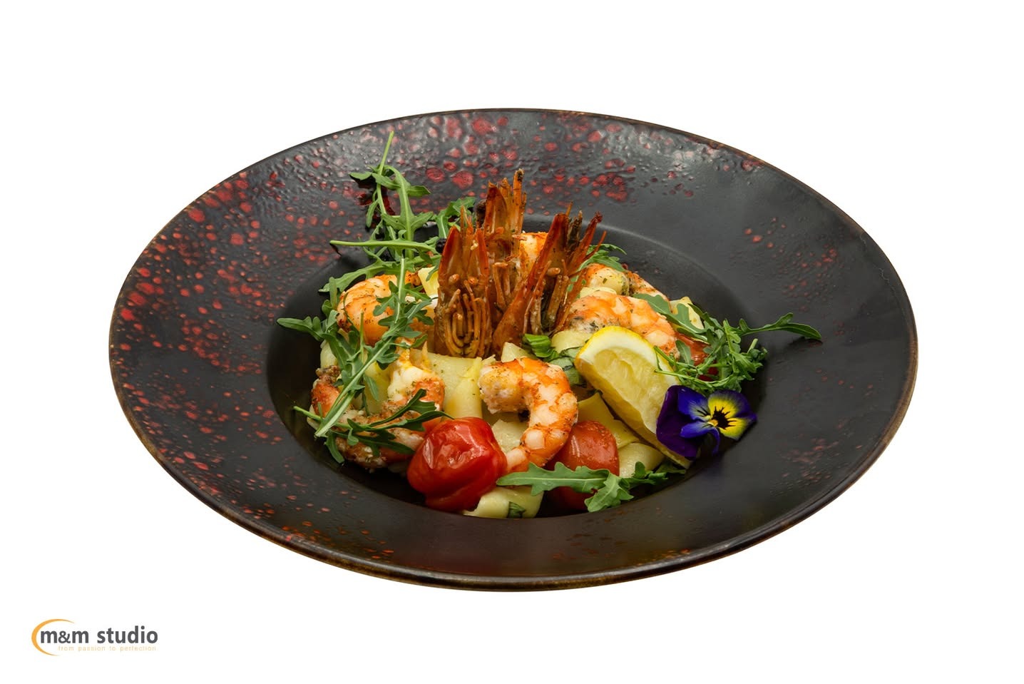 Kreatywne ujęcie makaronu z krewetkami na talerzu z ciemnej ceramiki, udekorowanego rukolą, pomidorkami, cytryną i bratkiem. Fotografia kulinarna na białym tle.