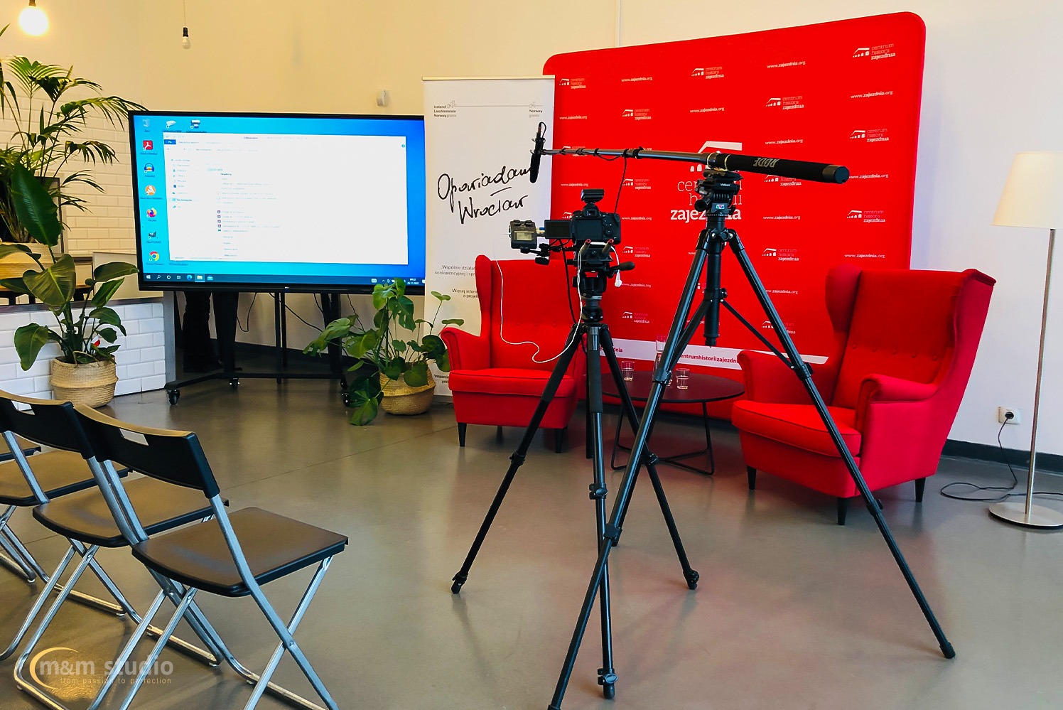 Kamera z mikrofonem na statywie w studio z czerwonymi fotelami i ekranem. Przygotowania do nagrania, oświetlenie studyjne, rośliny doniczkowe w tle.