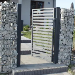 Nowoczesna brama ogrodzeniowa z poziomych profili, osadzona na słupkach z gabionów wypełnionych jasnymi kamieniami, prowadząca do domu z numerem 19.
