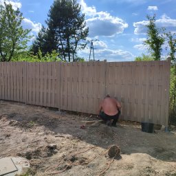 Profi Garden Łukasz Majewski - Montaż drewnianego ogrodzenia panelowego: mężczyzna w czarnych spodniach i bez koszulki pracuje przy mocowaniu paneli do słupków, widoczny kabel zasilający, narzędzia i wiadro, w tle drzewa i niebo