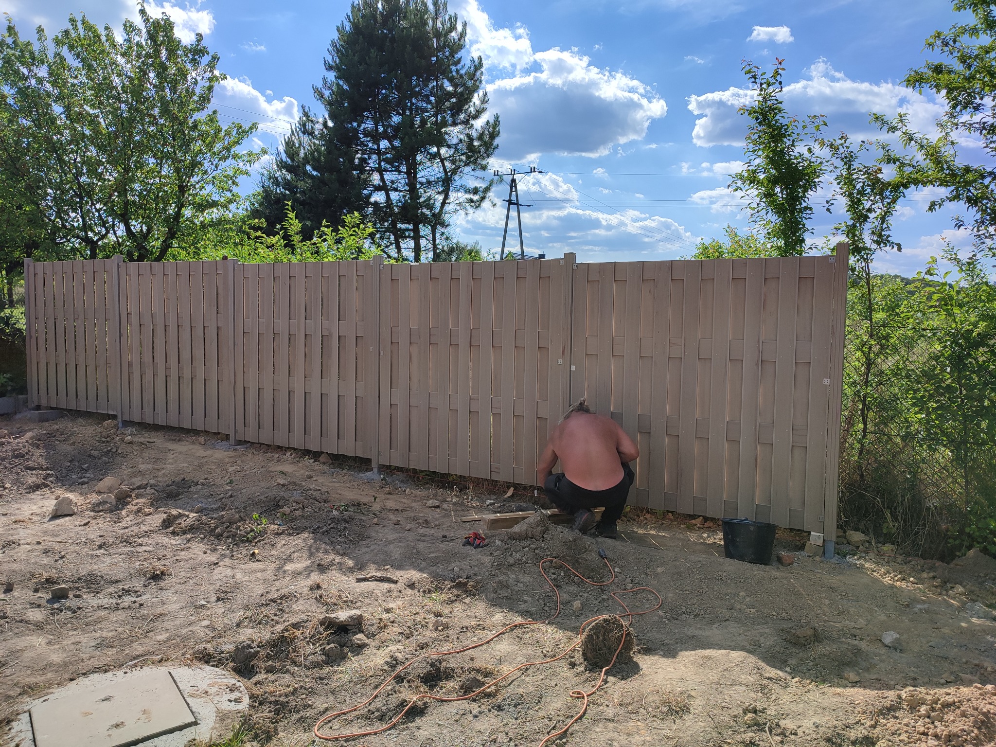Montaż drewnianego ogrodzenia panelowego: mężczyzna w czarnych spodniach i bez koszulki pracuje przy mocowaniu paneli do słupków, widoczny kabel zasilający, narzędzia i wiadro, w tle drzewa i niebo