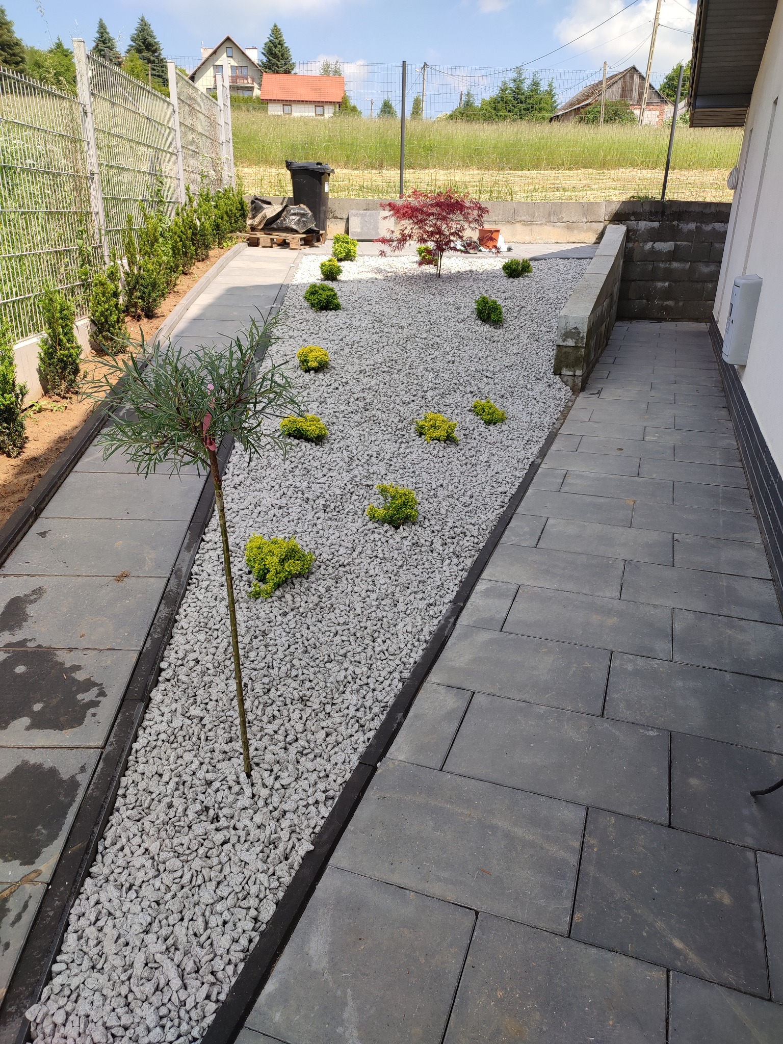 Nowoczesny ogród z szarymi kamieniami, ścieżką z płyt betonowych i ozdobnymi krzewami oraz drzewkami, widok z góry.