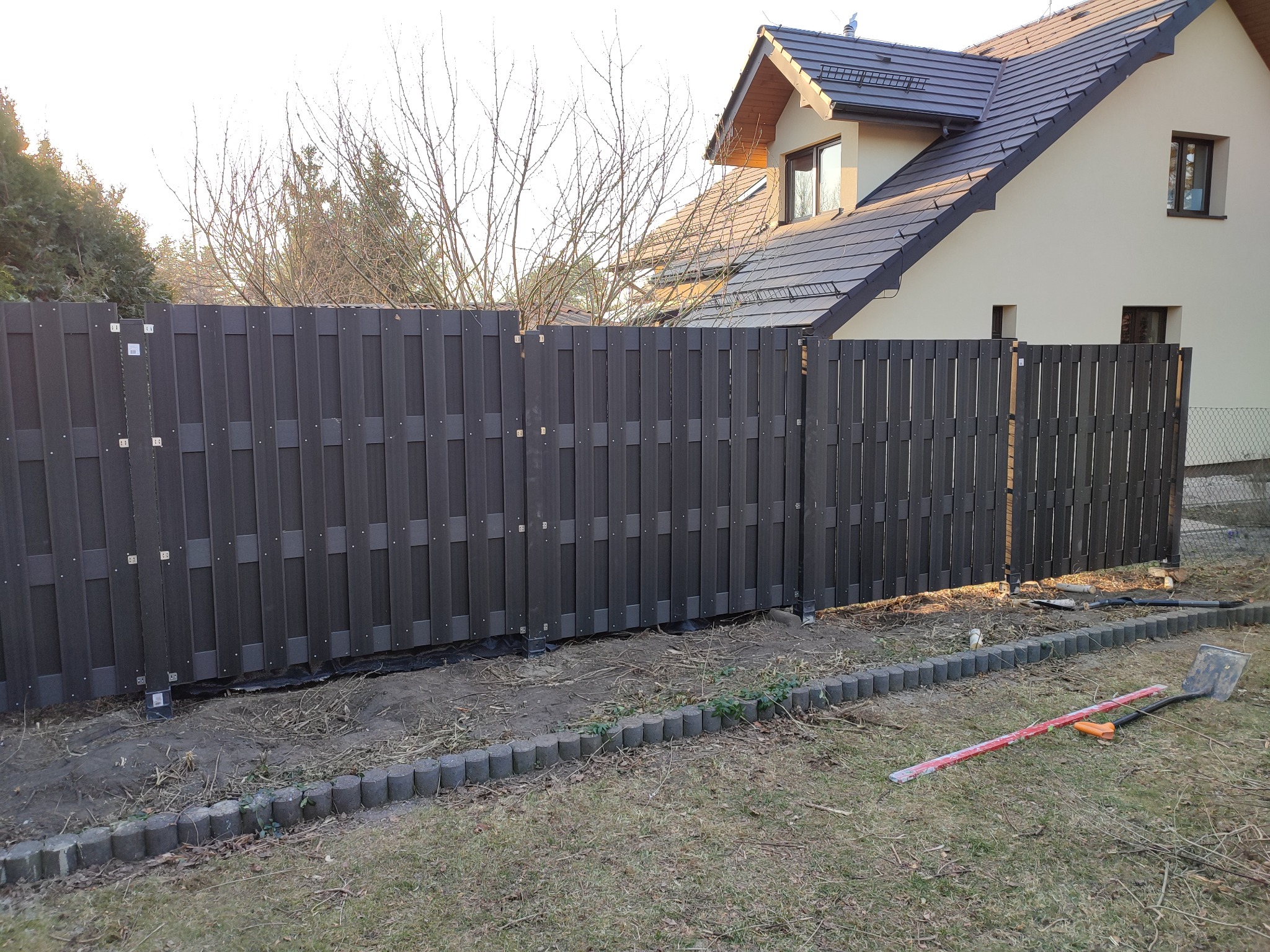 Nowo postawione, ciemnobrązowe, panelowe ogrodzenie z tworzywa sztucznego na tle jasnego domu z ciemnym dachem. W tle drzewa bez liści. Na pierwszym planie trawa, obrzeża z kostki i narzędzia...
