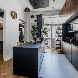 Nowoczesna kuchnia z czarną wyspą i drewnianymi szafkami, oświetlona wiszącymi lampami, z widokiem na balkon i salon z ceglaną ścianą, dekorowana roślinami doniczkowymi i złotym zegarem na ścianie.