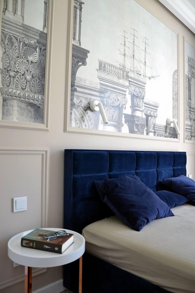 Elegancka sypialnia z granatowym, aksamitnym zagłówkiem, nocnym stolikiem z książką i okularami, oraz dekoracyjnymi panelami ściennymi z motywem architektonicznym i oświetleniem punktowym.