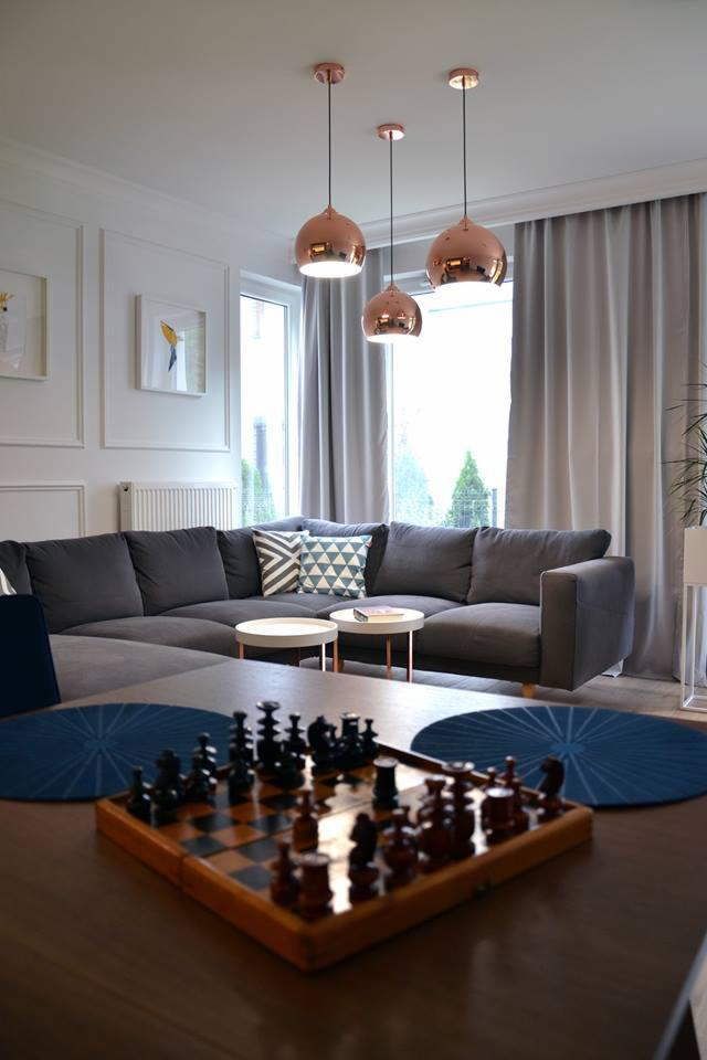 Jasne wnętrze salonu z szarym narożnikiem, okrągłymi stolikami kawowymi z miedzianymi nogami, miedzianymi lampami wiszącymi i szachami na pierwszym planie.