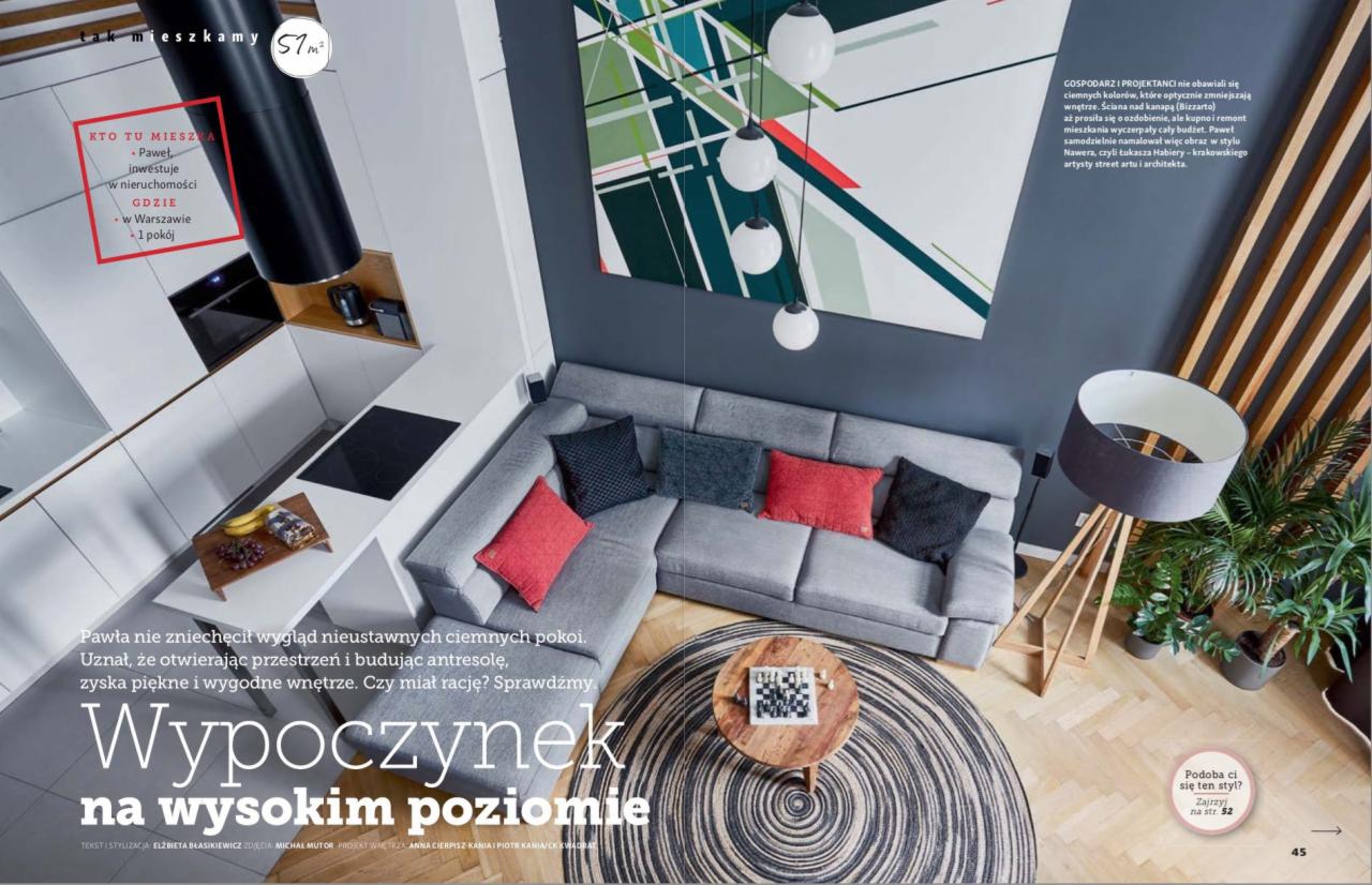 Zdjęcie z góry przedstawia nowoczesne mieszkanie z szarą sofą narożną, okrągłym dywanem w paski, drewnianym stolikiem kawowym z szachami, białymi szafkami kuchennymi i abstrakcyjnym obrazem...