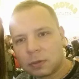 Portret mężczyzny z krótkimi włosami, widoczny fragment szyi z łańcuszkiem, w tle osoby i napis NOVA