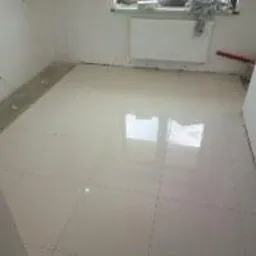 Błyszcząca, jasna podłoga wyłożona kwadratowymi płytkami w pustym pomieszczeniu, widoczne fugi i odbicie światła, przygotowane do dalszych prac wykończeniowych.