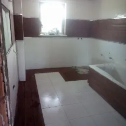 Łazienka w trakcie remontu: białe płytki podłogowe częściowo ułożone, brązowe panele ścienne na górze, wanna zabezpieczona folią, widoczne niedokończone prace przy podłodze i ościeżnicy.