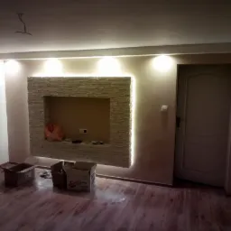 Ściana z dekoracyjną wnęką wykończoną kamieniem elewacyjnym z podświetleniem LED, widoczne kartony na podłodze, drewnopodobne panele podłogowe i białe drzwi w jasnym wnętrzu.